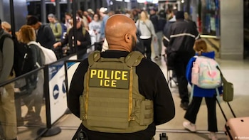 Vista trasera de un hombre calvo con barba, vestido con un chaleco verde oliva con las letras "POLICE ICE" en amarillo, de pie en un concurrido pasillo con gente