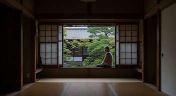 Interior de una casa japonesa con suelo de tatami y paneles shoji; una persona adulta mayor se sienta en una ventana abierta que da a un jardín tradicional.