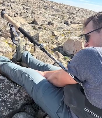 Un hombre acostado en el suelo rocoso con pantalones azules y una camiseta gris. Un bastón de trekking con empuñadura de madera está apoyado junto a él