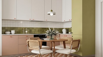 Una cocina con gabinetes blancos y rosas, un mueble alto verde, y un salpicadero de azulejos verdes. Mesa redonda de madera con cuatro sillas de ratán y tela blanca.