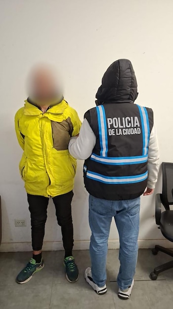 Un efectivo con chaleco de la Policía de la Ciudad sostiene a un hombre con chaqueta amarilla y rostro difuminado de pie ante una pared blanca