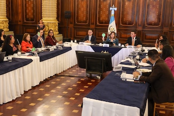 Veinte personas en un salón con paredes de madera y columnas doradas. Mesas largas con manteles. Bandera de Guatemala. Dos personas sostienen tarjetas de votación