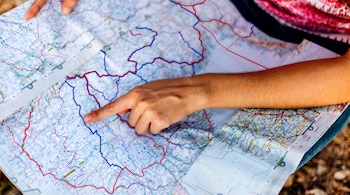 Dos manos sostienen un mapa de papel extendido con rutas, ciudades y fronteras; una mano con el dedo índice señala un punto en el mapa.