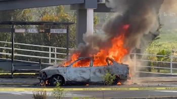 Vehículo de color claro en llamas con fuego y humo negro, estacionado en una calle. Se observa cinta de precaución amarilla, vegetación y señales viales borrosas.
