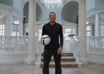 Kylian Mbappé, un hombre joven sonriente, de pie en un vestíbulo blanco con columnas y pasamanos ornamentados, sostiene un balón de fútbol