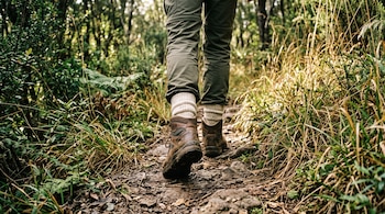 Vista en ángulo bajo de las piernas y botas de senderismo de una persona caminando por un camino de tierra con vegetación.