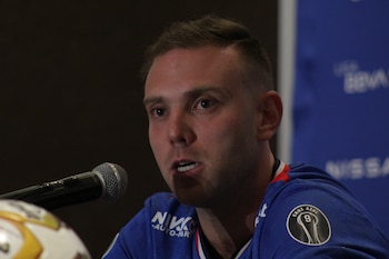 Primer plano de Joel Huiqui en una conferencia, con un micrófono plateado al frente y vistiendo una camiseta azul de Cruz Azul con un escudo