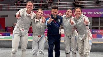 Cuatro mujeres vestidas con uniforme de esgrima y un hombre con chaqueta azul posan sonrientes en una pista deportiva.