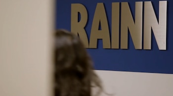 Letras metálicas con la palabra RAINN fijadas en una pared azul, junto a la figura desenfocada de una persona