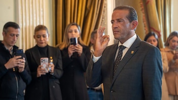 Un hombre con traje gris levanta su mano derecha mientras tres personas en el fondo lo fotografían con teléfonos móviles