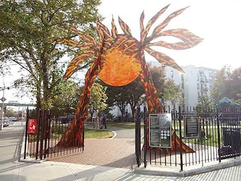 Escultura colorida con forma de sol instalada sobre la entrada de una cerca de metal negro en un parque urbano con árboles y edificios al fondo