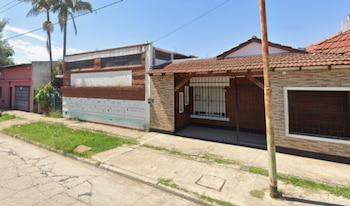 Exterior de un edificio de ladrillo con techo de tejas y ventanas enrejadas. Se ve una acera, vegetación y palmeras bajo un cielo azul