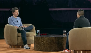 Dos hombres sentados en sillones frente a una mesa de café con vasos de agua. El hombre de la izquierda viste camiseta azul y zapatillas blancas. El otro, de espaldas, usa camiseta oscura