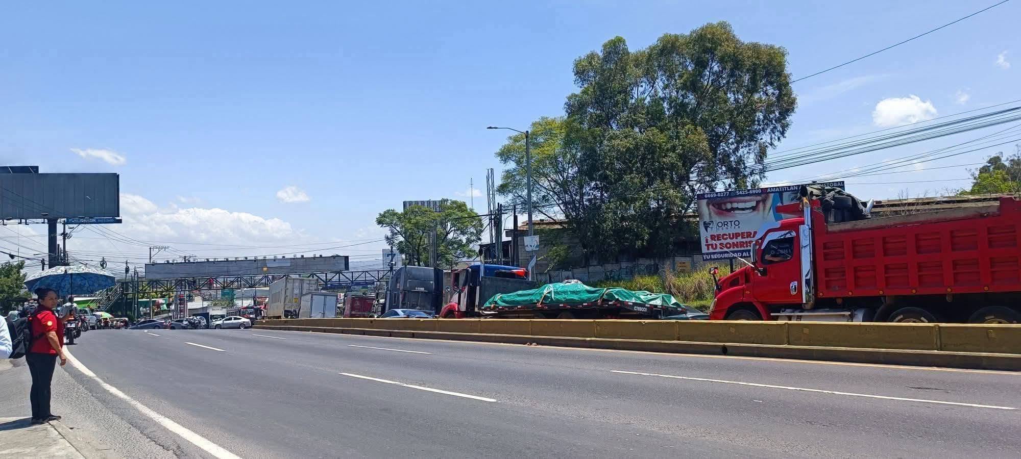 Varios camiones de carga y automóviles transitan por una carretera urbana bordeada por vallas publicitarias y árboles en un día despejado. (Redes Sociales)