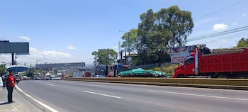 Camiones de carga transitan por una carretera asfaltada con vallas publicitarias al fondo y una mujer con sombrilla a la izquierda