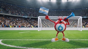 Una mascota roja con el número 6 en el centro sostiene una bandera argentina y una vuvuzela azul en un estadio de fútbol con gradas llenas.