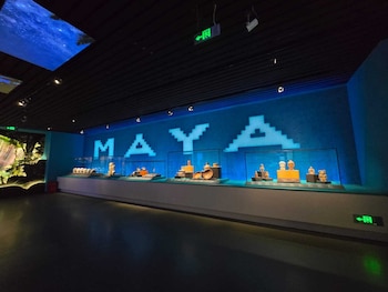Sala de museo con vitrinas que contienen figuras y vasijas de cerámica. Letrero luminoso 'MAYA' en pared azul. Techo oscuro con luces, pantallas en el cielo y un mural de selva en la pared izquierda. Cartel de salida verde