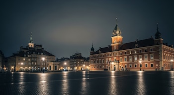 Vista nocturna de una plaza adoquinada con edificios históricos iluminados a su alrededor, incluyendo un castillo o palacio con torres. El suelo mojado refleja las luces.