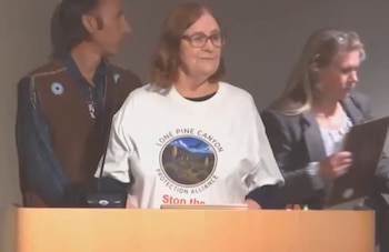 Una mujer con gafas y camiseta blanca con la leyenda Lone Pine Canyon Protection Alliance está detrás de un podio de madera