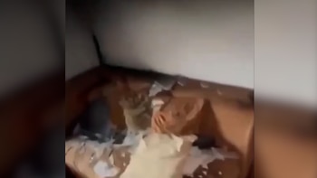 Interior de una habitación dañada, con paredes y techo ennegrecidos. Hay objetos quemados y escombros dispersos sobre lo que parece ser un sofá