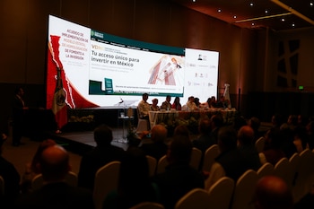 Público y oradores en evento, pantalla grande con sitio web 'Gobierno de México' y texto sobre inversiones y Baja California, bandera mexicana