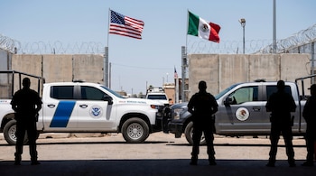Una camioneta blanca de la Patrulla Fronteriza de EE. UU. y una gris mexicana estacionadas frente a un muro fronterizo, con siluetas de agentes, barreras y banderas.