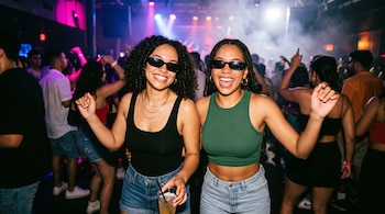 Dos jóvenes con gafas oscuras y camisetas sin mangas sonríen en la pista iluminada de una discoteca repleta de gente.