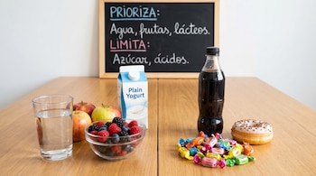 Mesa con opciones de alimentos contrastadas: agua, manzanas, frutos rojos y yogur (izquierda), y refresco, caramelos y una dona (derecha). Una pizarra al fondo detalla recomendaciones.