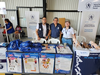 Dos trabajadoras con chalecos azules están tras una mesa con jeringas, recipientes médicos y una pancarta sobre vacunación, peso y talla