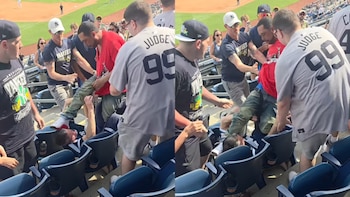 Múltiples aficionados participan en una pelea entre los asientos de un estadio de béisbol. Un hombre está en el suelo mientras otros están de pie alrededor