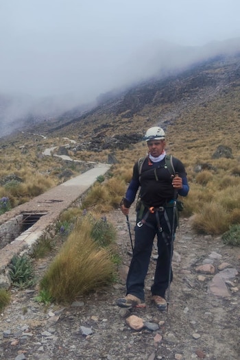 Un hombre con casco, mochila y bastones de trekking se encuentra de pie en un camino rocoso de montaña cubierto por niebla