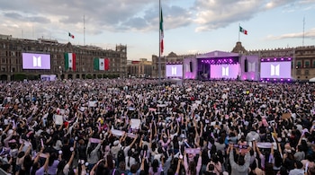 Vista elevada de una multitud masiva en el Zócalo de Ciudad de México, con gente alzando luces y pancartas. Se ven pantallas con el logo de BTS y banderas mexicanas.