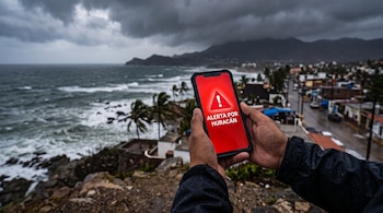 Unas manos sostienen un celular con pantalla roja que dice Alerta por huracán frente a una costa con olas y palmeras.