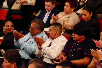 Un grupo de personas sentadas en un auditorio aplaude. Hombres y mujeres con vestimenta de patrones coloridos. Sillas rojas visibles