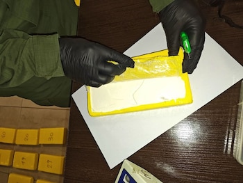 Manos enguantadas de un oficial abriendo un paquete rectangular envuelto en plástico amarillo que contiene una sustancia blanca, sobre una mesa de madera