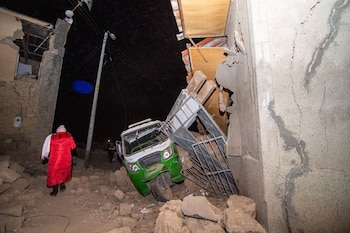 Vivienda con pared derrumbada y escombros en el suelo, un mototaxi verde y blanco aplastado, una persona con abrigo rojo caminando de espaldas