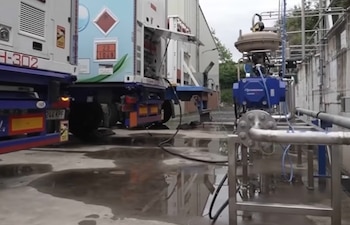 Un camión cisterna blanco y azul con matrículas y señalización de peligro estacionado en un área industrial, conectado a equipos de bombeo y tuberías; el suelo está mojado