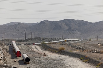 Tuberías metálicas en el suelo, un camino de tierra, postes con cables eléctricos y montañas rocosas al fondo