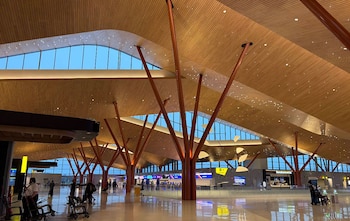 Vista interior de un moderno terminal de aeropuerto con altas columnas de madera, techo ondulado iluminado y grandes ventanales. Personas y carros de equipaje en el suelo brillante