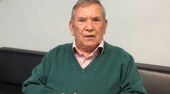 Un hombre mayor con suéter verde y camisa rosada mira hacia el frente sentado junto a un sillón oscuro.