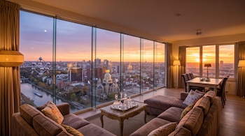 Interior de un salón amueblado con ventanales que dan a una vista urbana de Bruselas con el Atomium al fondo durante el atardecer.