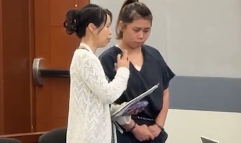 Dos mujeres de pie. Una lleva uniforme oscuro y la otra un cárdigan claro con un portapapeles. Fondo con puerta de madera y una pared clara