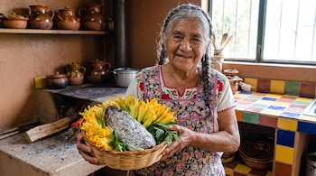 Mujer de edad con trenzas y delantal bordado sostiene una canasta con elote con huitlacoche, flores de calabaza y hojas verdes en una cocina rústica.