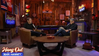 Dos hombres sentados en sillones con micrófonos frente a una mesa de café, en un estudio de televisión con un billar y decoraciones en las paredes