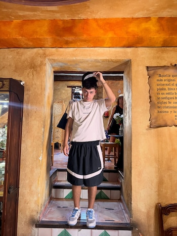 Un joven, Trueno, de pie en un arco con paredes ocres, vistiendo camiseta blanca, shorts negros y zapatillas claras, ajustándose una gorra