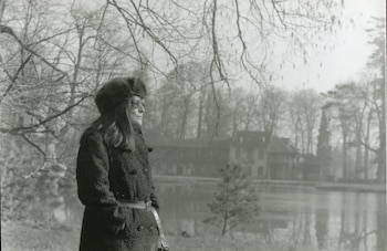 Fotografía en blanco y negro de Mercedes Sosa con gorro de piel, abrigo y gafas, de pie al lado de un lago con árboles sin hojas y un edificio en el fondo