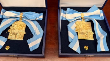 Dos medallas doradas con cintas celestes y blancas en cajas azules. Las medallas tienen la inscripción Medio Ambiente 2023