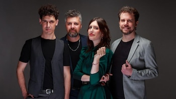 Cuatro músicos posan en estudio. Tres hombres y una mujer de pie, sobre un fondo gris. La mujer está en el centro, los hombres a sus lados