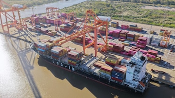 Vista aérea de un buque de carga negro cargado con contenedores coloridos en un puerto con grúas naranjas y pilas de contenedores en el muelle