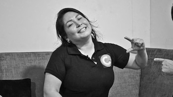 Fotografía en blanco y negro de Susy Aponte Polo sonriendo en un sofá, vestida con un polo oscuro con un distintivo circular